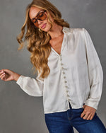Woman wearing a cream satin button-front blouse with V-neckline and long sleeves, styled with dark denim jeans and aviator sunglasses