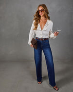 Woman in a cream satin button-front blouse, dark wide-leg jeans, brown belt, aviator sunglasses, woven clutch and heeled sandals