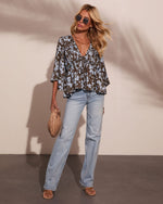 Woman wearing olive floral boho blouse with bell sleeves, light wash wide-leg jeans, strappy sandals, and woven clutch