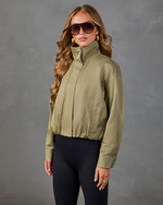 Woman wearing olive green zip-up bomber jacket with mock neck and black leggings, styled with gold earrings and oversized sunglasses