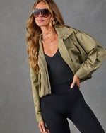 Woman wearing olive green cropped bomber jacket, black bodysuit, and leggings with aviator sunglasses and layered gold necklaces