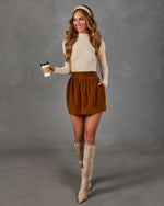 A woman modeling a cream ribbed long-sleeve top and a brown corduroy mini skirt, paired with beige knee-high boots, holding a coffee cup against a gray studio backdrop.