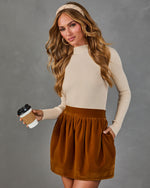 A woman modeling a cream ribbed mock-neck long-sleeve top paired with a brown corduroy skirt, holding a coffee cup against a gray studio background.