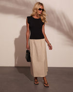 Woman wearing black sleeveless top with padded shoulders and beige maxi skirt, black sandals, sunglasses, and woven bag