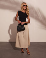 Woman in black sleeveless top, beige midi skirt, black sandals, holding woven leather handbag with gold hardware