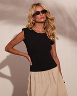 Woman wearing black sleeveless knit top with shoulder detail and beige midi skirt, styled with small black sunglasses and gold earrings