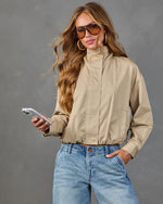 Woman wearing cropped khaki zip-up jacket and light wash high-waist jeans, holding smartphone, aviator sunglasses, gold hoops