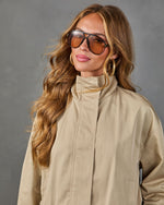 Woman wearing beige trench coat, oversized brown aviator sunglasses, gold hoop earrings, and wavy blonde hair against gray background