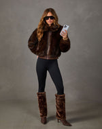 Woman wearing a brown faux fur bomber jacket, black leggings and fur foldover heeled boots while holding a smartphone