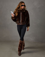 Woman in brown faux fur bomber jacket, black leggings, fur foldover boots and aviator sunglasses holding a phone