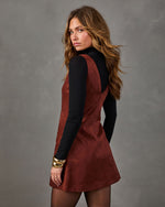 Woman in a rust suede mini pinafore dress layered over a black turtleneck, with sheer tights and chunky gold bracelets
