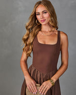 Woman in chocolate brown square-neck sleeveless midi dress with gathered skirt, gold jewelry, holding woven clutch