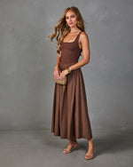 Brown midi dress with scoop neck bodice and flowy skirt, styled with strappy heels and woven clutch on female model