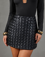 A close-up of a model wearing a black woven faux-leather mini skirt paired with a black long-sleeve top and gold cuff bracelets, set against a solid gray studio background.