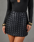 A close-up of a model wearing a black woven faux-leather mini skirt paired with a black long-sleeve top and gold cuff bracelets, set against a solid gray studio background.