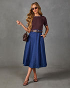 A woman modeling a short-sleeve crewneck top in dark plum with white piping trim, paired with a blue denim midi skirt, a brown belt, sunglasses, and strappy heels against a gray studio backdrop.
