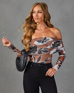 Woman wearing brown floral off-shoulder mesh bodysuit with long sleeves, black high-waist jeans, and woven black handbag