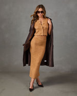 A model wearing a camel-colored knit two-piece set consisting of a sleeveless button-front top and a matching pencil skirt, styled with a dark brown textured coat draped over her shoulders, sunglasses, and dark brown pointed-toe heels against a gray studio backdrop.