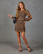 A woman modeling a long-sleeve mini dress with a brown and black floral lace pattern, paired with black strappy heels and a small black clutch, against a gray studio backdrop.