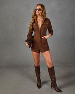 A woman models a brown faux-suede jumpsuit with a front zipper and chest pockets, paired with matching brown knee-high boots and sunglasses, against a gray studio backdrop.