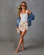 Woman wearing white bodysuit, floral mini skort, denim jacket, strappy heels, and sunglasses in a summer fashion editorial look.