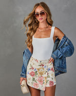 Woman wearing white bodysuit, floral mini skirt, denim jacket, aviator sunglasses, layered necklaces, holding cream crossbody bag