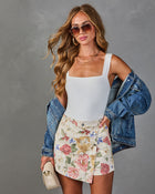 Woman wearing white bodysuit, floral mini skirt, denim jacket, aviator sunglasses, layered necklaces, holding cream crossbody bag
