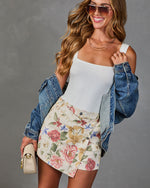 Woman wearing white bodysuit, floral mini skirt, denim jacket, aviator sunglasses, holding cream crossbody bag