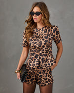 A woman modeling a matching leopard-print short-sleeve top and shorts set against a textured gray studio background. She is wearing black sunglasses, gold hoop earrings, a gold bracelet, and holding a black clutch.