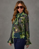 Woman in sheer green floral burnout velvet blouse with neck bow and bell sleeves, styled with aviator sunglasses and dark jeans