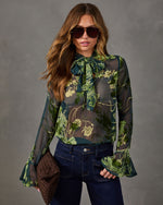 Sheer green floral velvet burnout blouse with neck bow and bell sleeves, styled with dark wide-leg jeans and a woven clutch