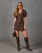 A woman modeling a brown short-sleeve mini dress with puff sleeves, paired with matching brown knee-high boots, sunglasses, and a small brown handbag, against a textured gray studio background.