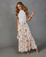 Woman wearing white sleeveless mock-neck top and cream floral maxi skirt with pink roses, gold bangles, nude heels