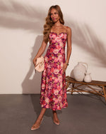 Woman wearing purple floral midi dress with spaghetti straps, holding beige clutch, styled with gold jewelry and clear heels