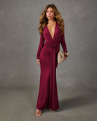 Burgundy long-sleeve maxi dress with plunging V-neckline and knotted waist, styled with gold jewelry and a woven beige clutch