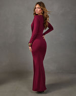 Woman in a burgundy long-sleeve maxi dress with draped bodice and gold cuff bracelets, posed from behind against a gray backdrop