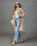 Woman modeling open tan trench coat over white ribbed top and blue jeans, wearing gray baseball cap, studio shot