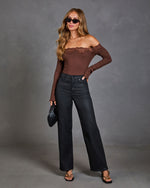 A woman models a brown off-the-shoulder ribbed knit top with a lace neckline, paired with black wide-leg jeans, black strappy sandals, and a black clutch, set against a textured gray studio background.