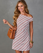 Woman wearing diagonal-striped one-shoulder mini dress in rose and white, holding woven tan bucket bag, gold bangles