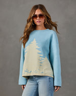 Woman in light blue oversized sweater with cream evergreen tree design, aviator sunglasses, gold hoops and light-wash jeans