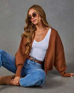 A woman wearing a brown open-front knit cardigan over a white ribbed tank top, blue jeans, and brown mule shoes, posed against a solid gray studio background.