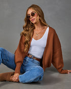 A woman wearing a brown open-front knit cardigan over a white ribbed tank top, blue jeans, and brown mule shoes, posed against a solid gray studio background.