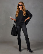 A woman modeling a black oversized batwing top, dark gray skinny leggings, and black over-the-knee boots, carrying a black studded tote bag against a textured gray studio backdrop.