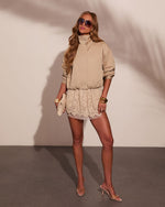Woman in beige bomber jacket, cream lace mini skirt, clear heeled mules, gold bangles, and oversized sunglasses holding a clutch.