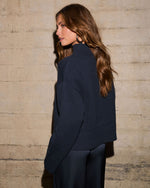 A woman wearing a dark navy blue ribbed knit mock-neck sweater, shown from a side-back angle against a textured concrete wall.