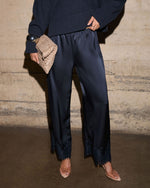 A model wearing dark navy wide-leg satin trousers with lace trim at the hem, paired with a dark navy sweater, beige pointed-toe heels, and holding a textured beige clutch against a concrete wall background.