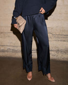 A model wearing dark navy wide-leg satin trousers with lace trim at the hem, paired with a dark navy sweater, beige pointed-toe heels, and holding a textured beige clutch against a concrete wall background.