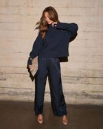 A woman modeling a dark navy blue ribbed knit sweater and matching wide-leg satin pants, holding a beige clutch against a textured concrete wall.