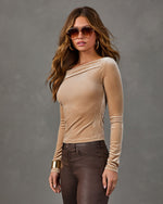 Woman wearing a beige velvet asymmetrical off-shoulder long sleeve top with brown coated skinny jeans, aviator sunglasses and gold jewelry
