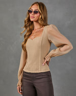 A woman modeling a beige ribbed long-sleeve top with sheer puff sleeves and a sweetheart neckline, paired with dark brown pants, against a textured gray studio background.